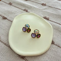 Darma Shop multi-stone stud earrings featuring faceted iridescent, purple, and emerald crystals in gold-toned settings.