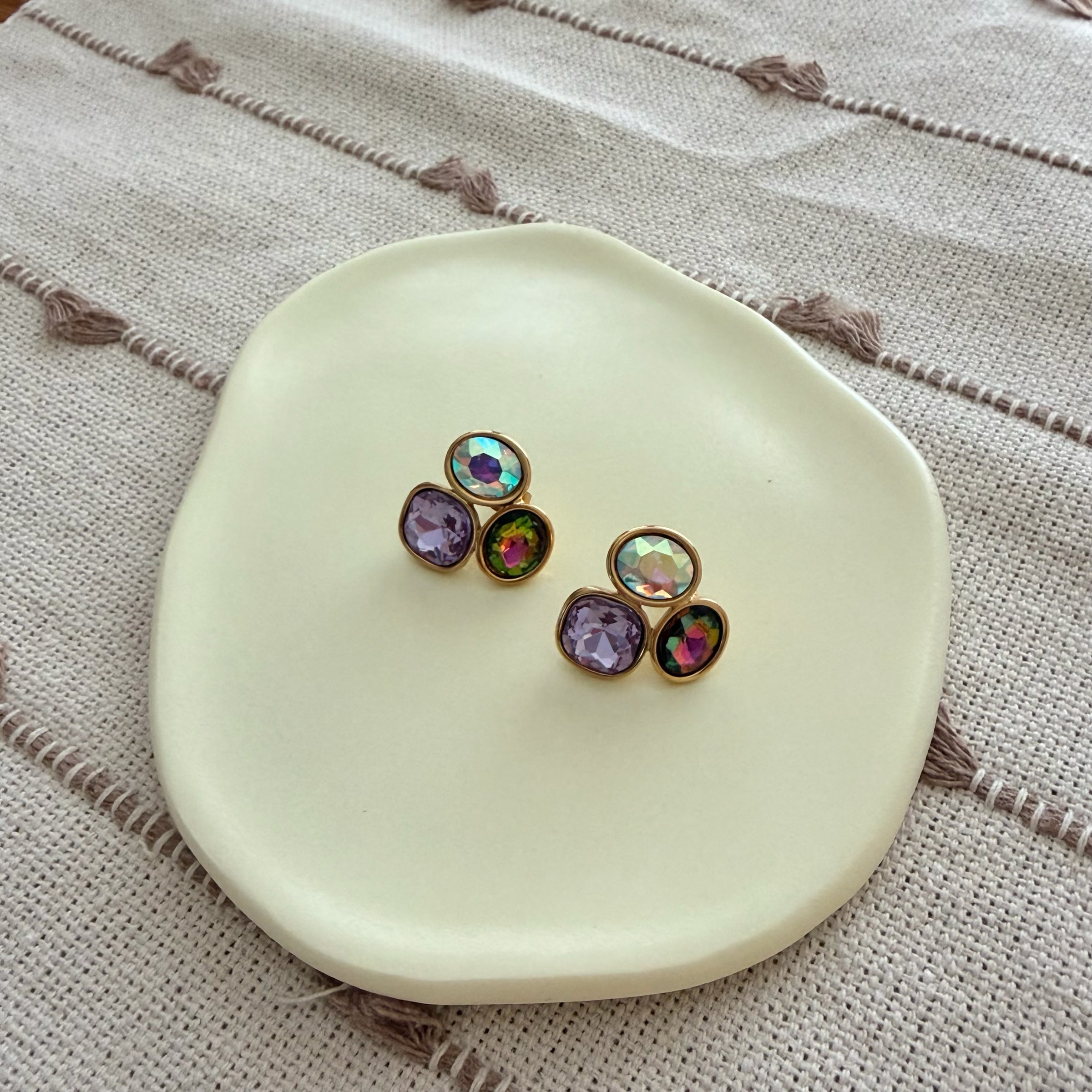 Darma Shop multi-stone stud earrings featuring faceted iridescent, purple, and emerald crystals in gold-toned settings.